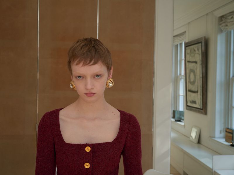 Close-up of a young woman with a pixie haircut wearing a square-necked dress and statement earrings, posing indoors.