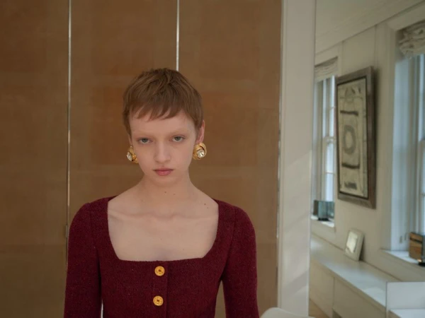Close-up of a young woman with a pixie haircut wearing a square-necked dress and statement earrings, posing indoors.