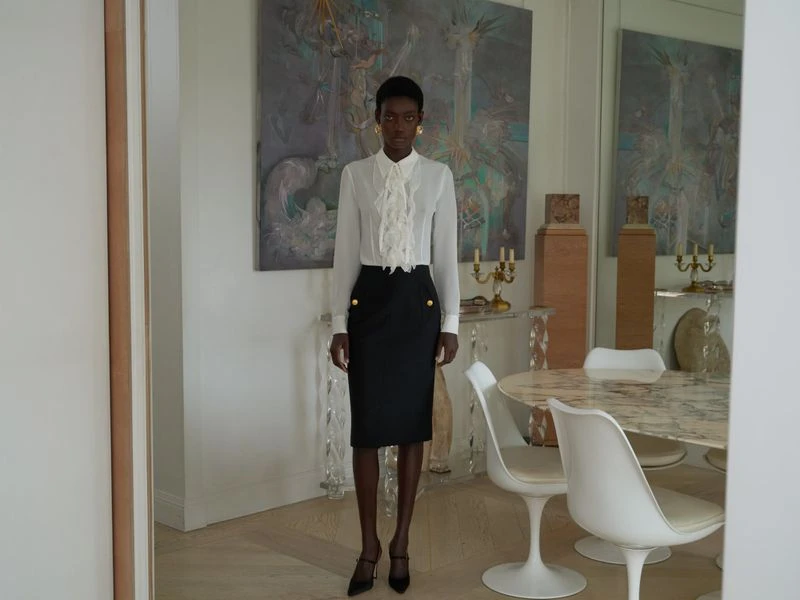 Elegant African American woman in white ruffled blouse and black skirt stands in a well-lit room with modern art and furniture.
