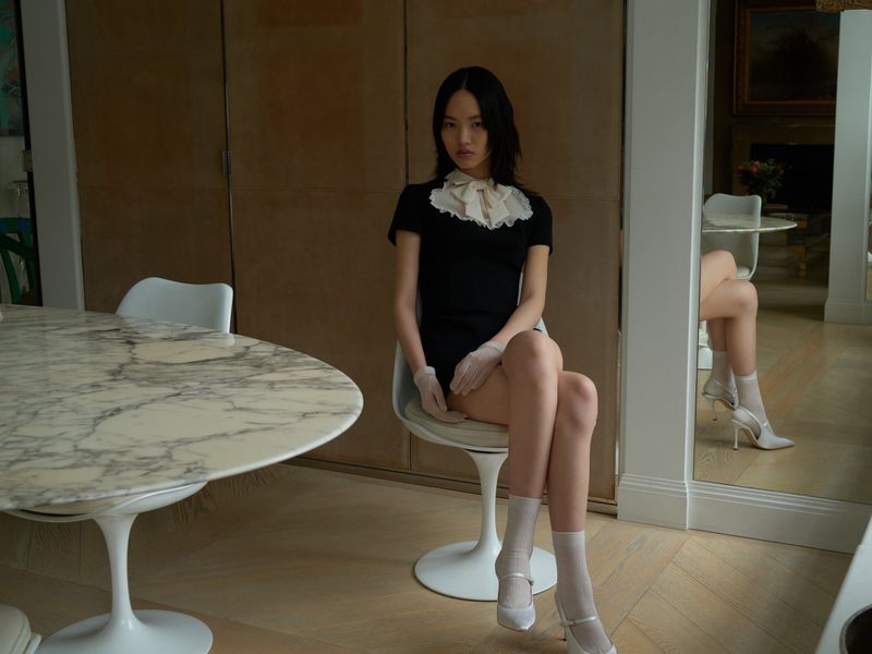 A young woman wearing a black mini dress with white lace collar and white gloves poses sitting on a chair in an indoor setting.