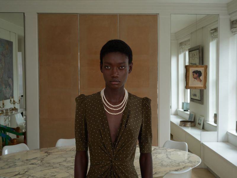 Elegant portrait of a Black woman in a polka-dot dress and pearl necklace, posing indoors with natural light in a neutral setting.