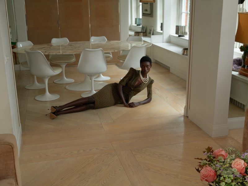 Elegant African American woman in patterned dress and heels poses on light wood floor in a bright interior near a floral arrangement.