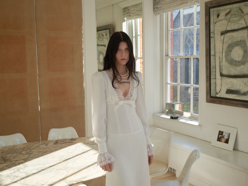 Young adult woman in a white dress with lace details, posing indoors with daylight, in an interior setting with a marble table.
