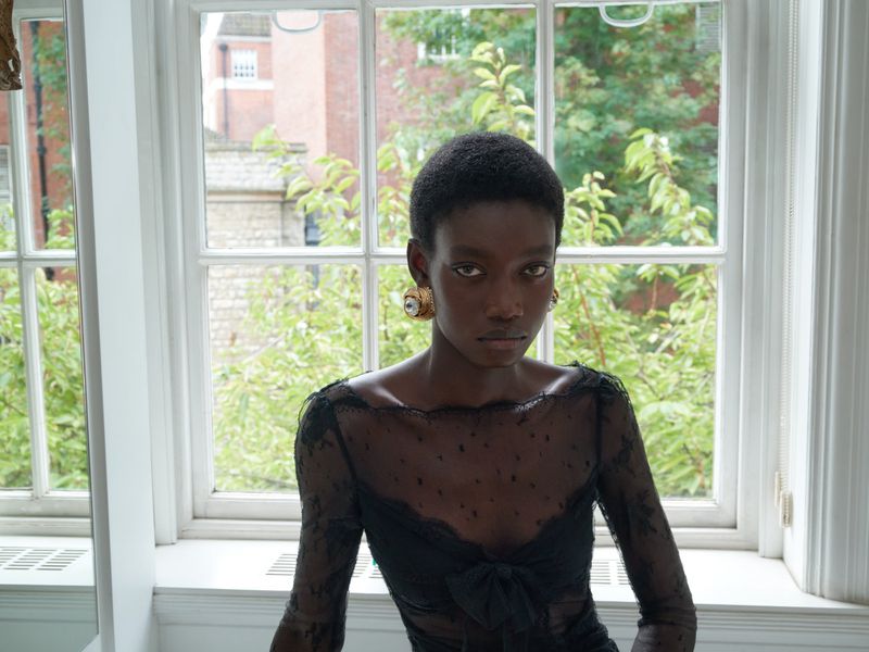 Close-up of an adult woman with short hair wearing a black lace top standing indoors near a window with outdoor greenery in the background.