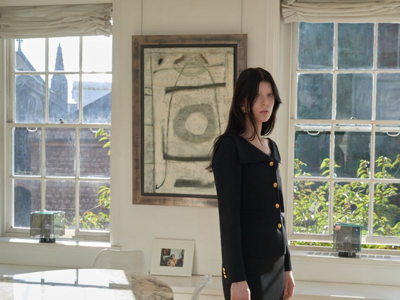 Indoor fashion shot of a woman in a black dress with gold buttons, standing near a window with an abstract painting on the wall.