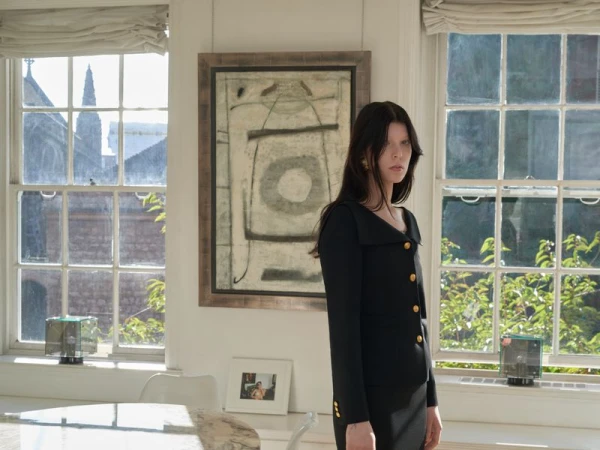 Indoor fashion shot of a woman in a black dress with gold buttons, standing near a window with an abstract painting on the wall.