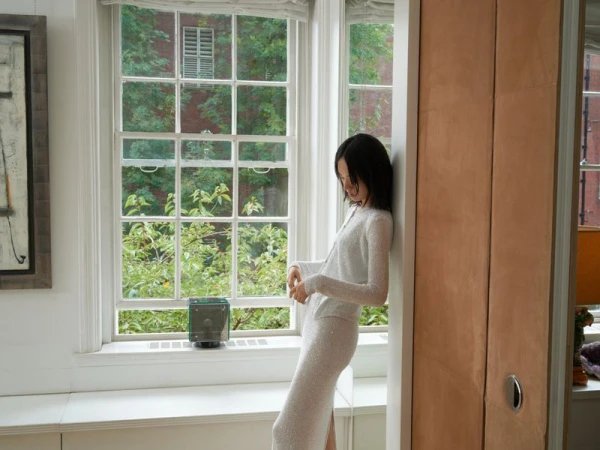 Indoor portrait of a woman in a sparkly white dress standing next to a window with trees and a glimpse of brick buildings outside.