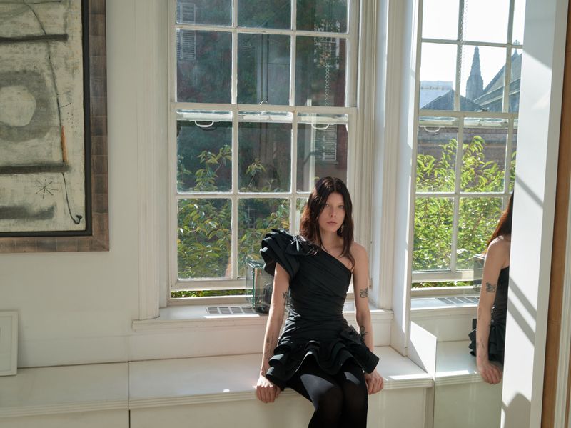 A woman in a black dress poses while sitting near a large window, with a painting and greenery visible outdoors.