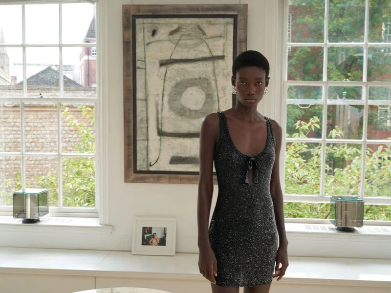 Indoor shot of a Black woman in a sparkly black mini dress in front of a large window and abstract painting.