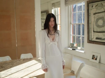 Model in a white dress with lace trim standing inside a bright room by a large window for Alessandra Rich Spring/Summer 2025.