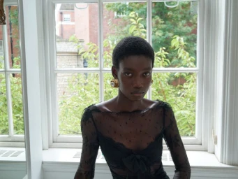 Model wearing black lace top and ornate earrings standing indoors before a large multi-pane window.