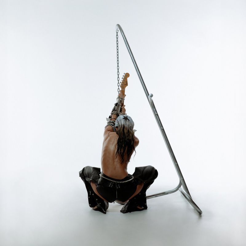 Back view of a bare-chested adult male squatting while suspended by chains attached to a guitar, set against a stark white backdrop.