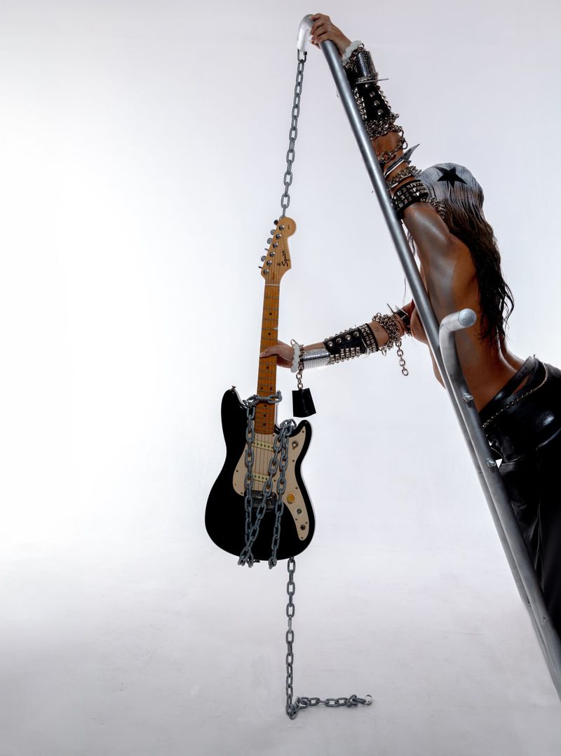 A slender adult figure poses with an electric guitar and a metal ladder, both draped with heavy metal chains, on a light background.