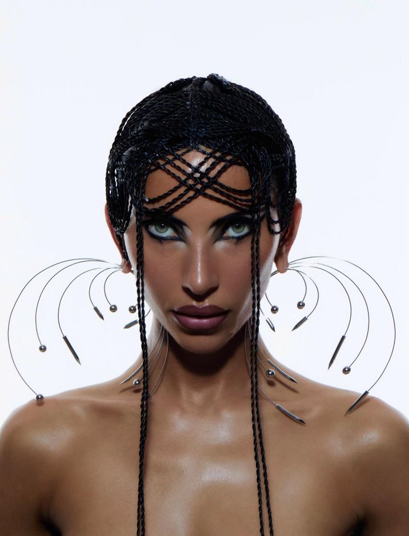 Close-up portrait of a woman with braided hair, dramatic makeup, and avant-garde earrings against a stark white background.