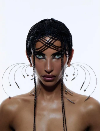 Close-up portrait of a woman with braided hair, dramatic makeup, and avant-garde earrings against a stark white background.