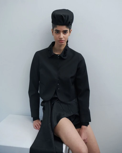 A woman poses wearing a black hat and matching tailored jacket, paired with a wrapped skirt, against a light grey studio backdrop.