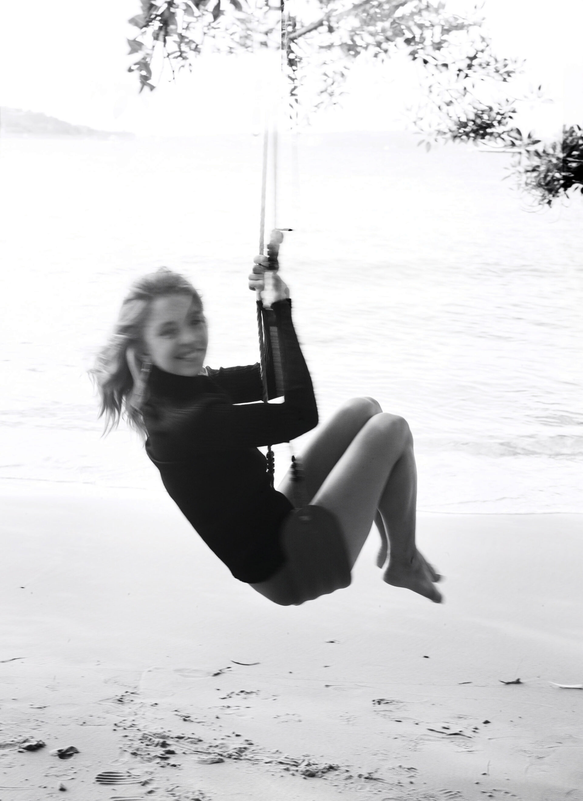 Monochrome photo of a woman swinging on a rope swing at a beach, with a blurred ocean and sky in the background.