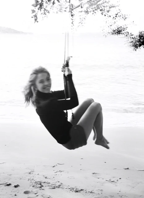 Monochrome photo of a woman swinging on a rope swing at a beach, with a blurred ocean and sky in the background.