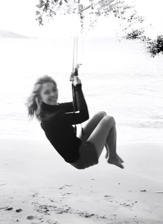 Monochrome photo of a woman swinging on a rope swing at a beach, with a blurred ocean and sky in the background.