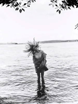 Sydney Sweeney standing in shallow water tossing her hair with foliage framing the top in a black and white editorial photograph.