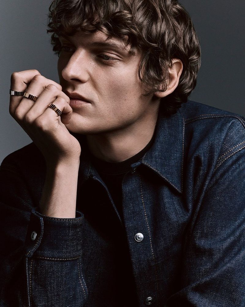 Man wearing a dark denim shirt displaying multiple gold rings on his fingers against a grey studio backdrop.