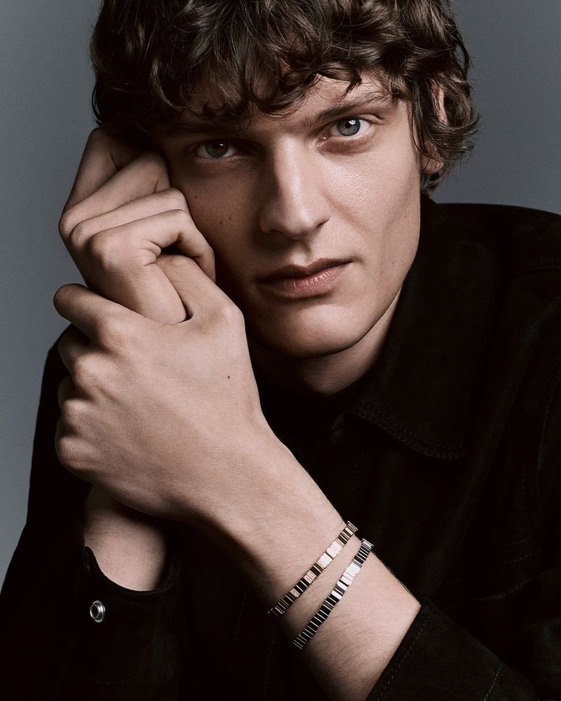 Portrait of a young adult male with curly brown hair wearing a black shirt and two bracelets against a grey background.