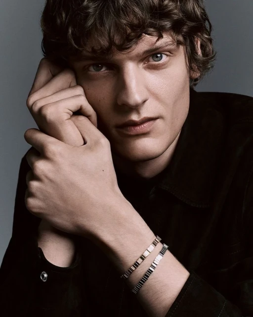 Portrait of a young adult male with curly brown hair wearing a black shirt and two bracelets against a grey background.