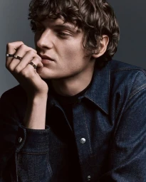 Man wearing a dark denim shirt displaying multiple gold rings on his fingers against a grey studio backdrop.