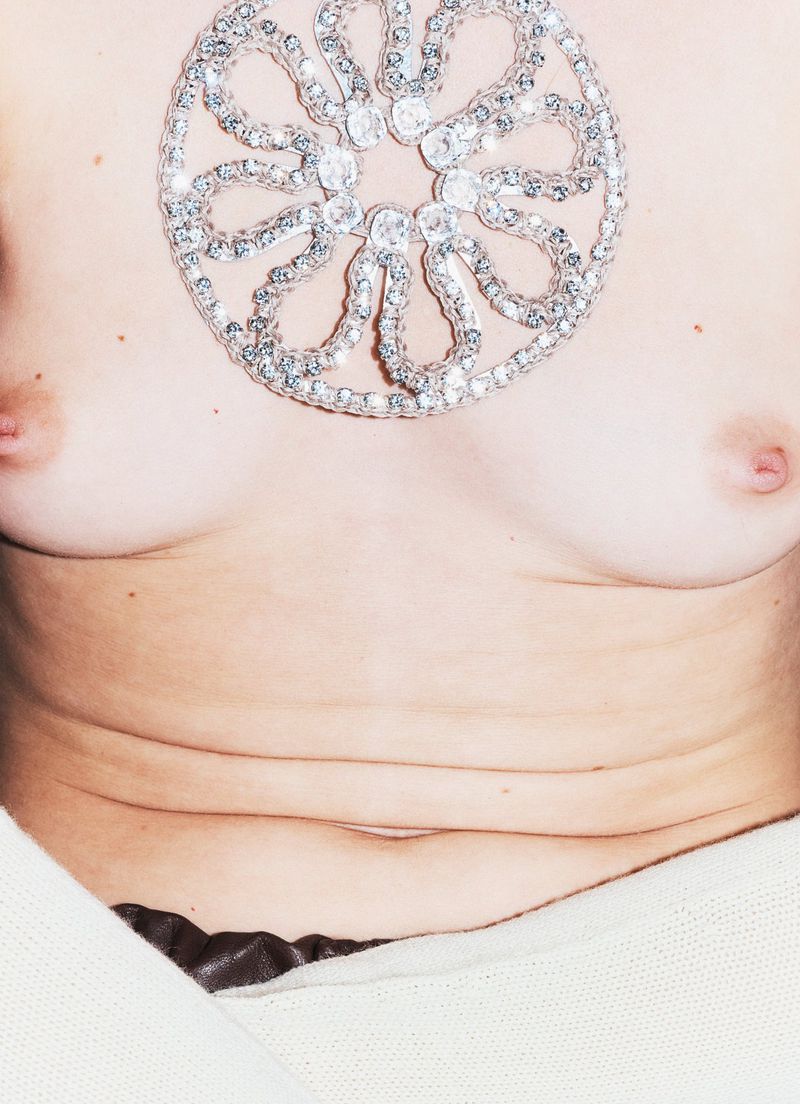 Close-up of a bare-chested person with a large, sparkly, circular embellishment; a neutral-toned blanket frames the bottom of the image.