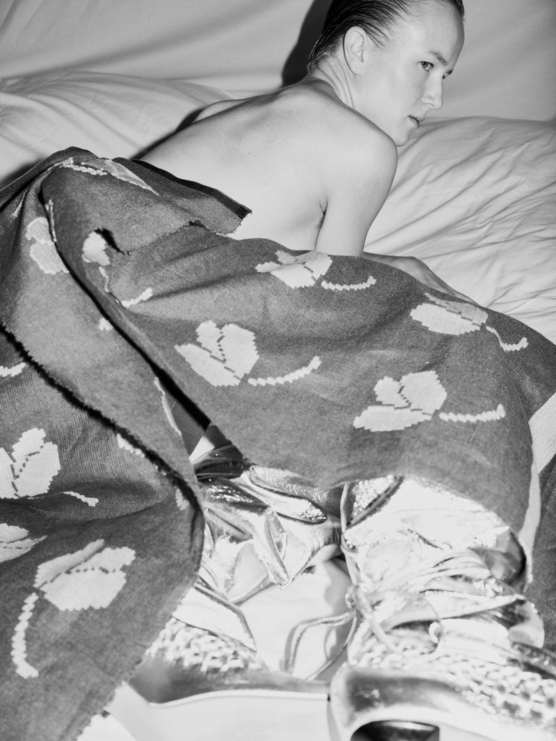 Model lying on a bed wrapped in a patterned blanket wearing metallic boots for Skarule Campaign S/S 25