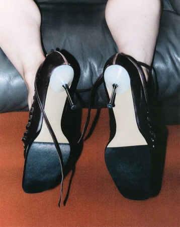 Close-up studio shot of a person wearing black high-heeled shoes with metal heels, seated on a brown surface with a black background.