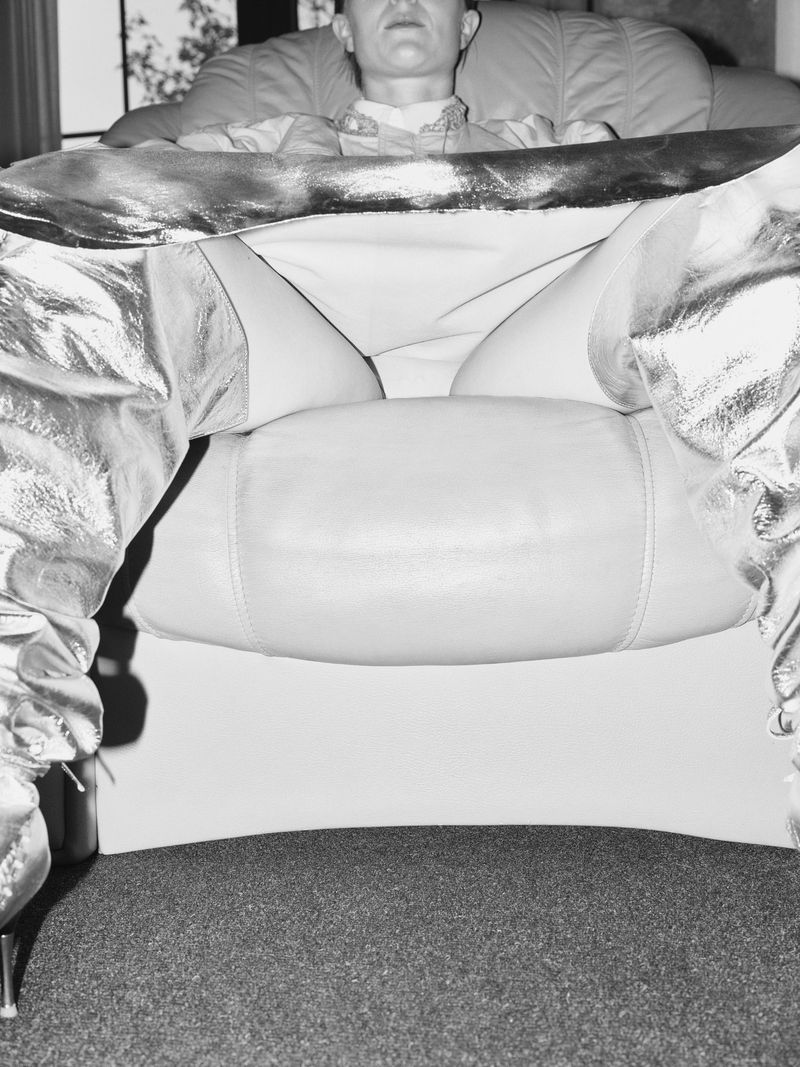 Black and white studio shot of a person lounging in a chair wearing shiny, metallic boots, capturing a unique perspective and style.