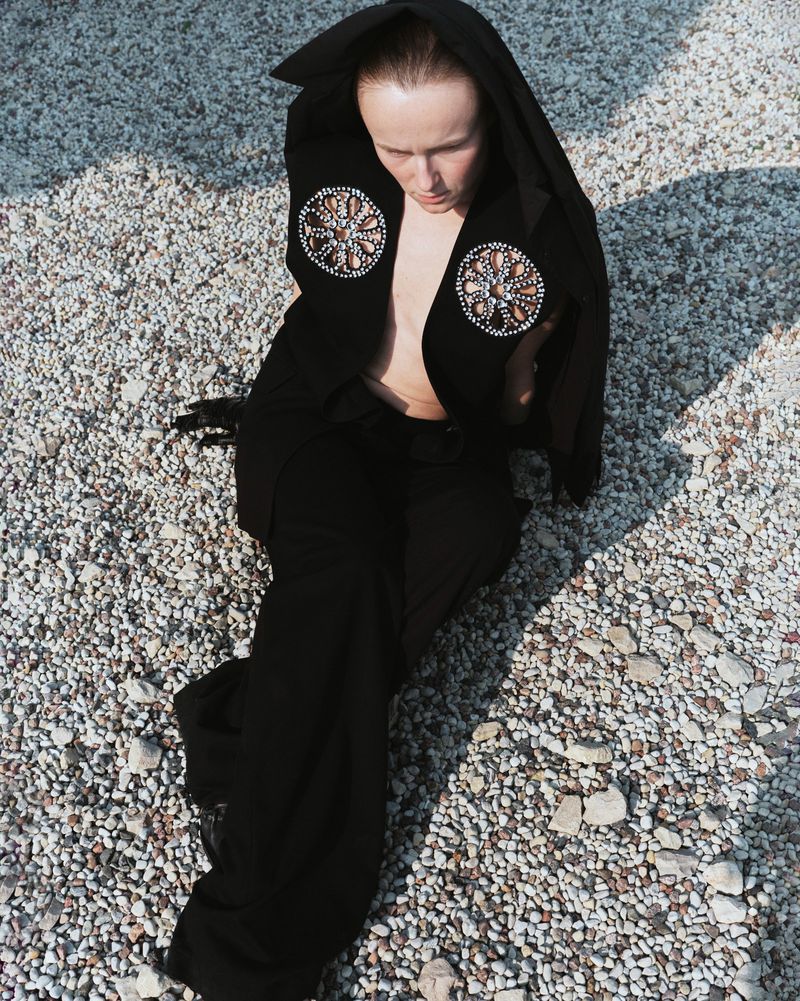Woman in black hooded jacket with circular beaded embellishments poses on a gravel surface in outdoor lighting, looking downward.
