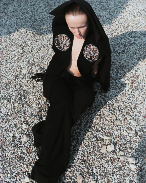 Woman in black hooded jacket with circular beaded embellishments poses on a gravel surface in outdoor lighting, looking downward.