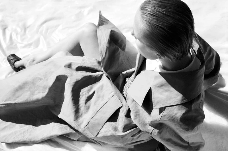 High-angle black and white shot of a person in a coat sitting on a white sheet, bathed in bright light, creating strong shadows.