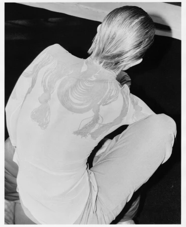 Monochrome shot from above of a person with blonde hair wearing a patterned shirt and light trousers, seated with their back to the viewer.