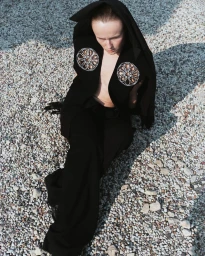 Model wearing a black hooded jacket with circular embellishments and black trousers seated on a gravel ground.
