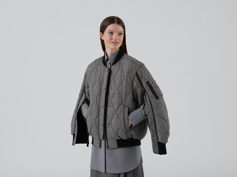 Studio shot of a smiling adult woman in a grey quilted bomber jacket, long grey skirt, and grey button-down shirt against a white background.