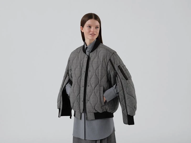 Studio shot of a smiling adult woman in a grey quilted bomber jacket, long grey skirt, and grey button-down shirt against a white background.