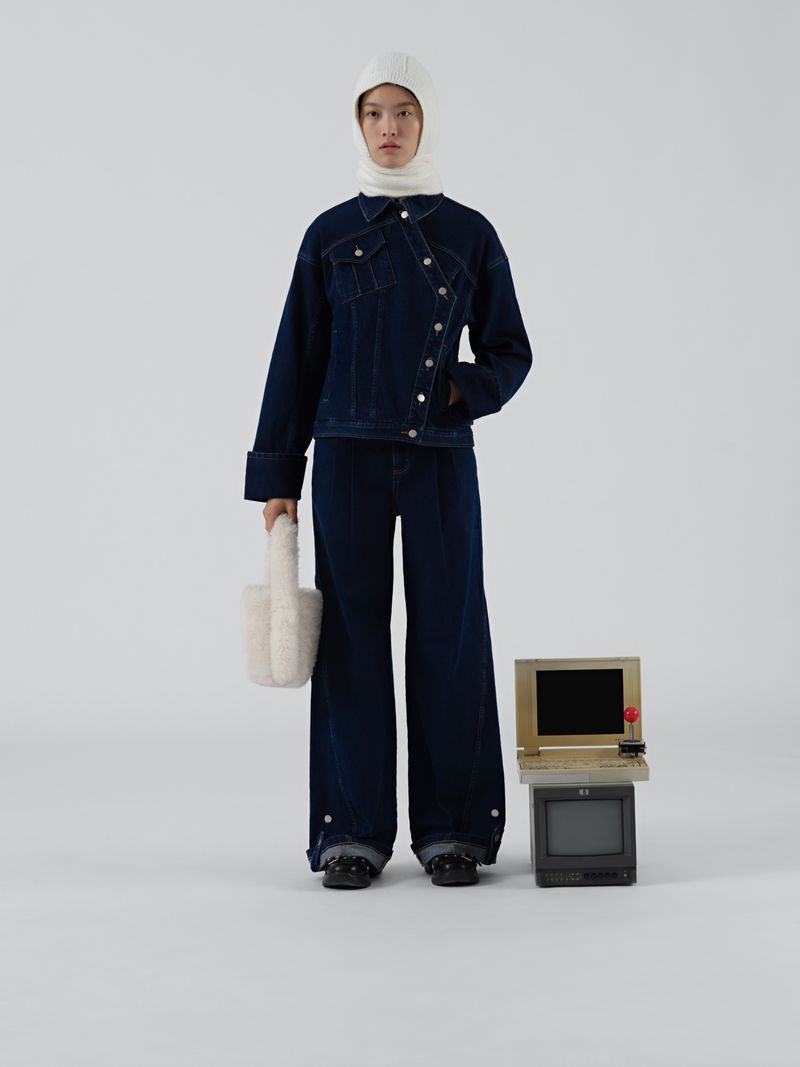 Woman in denim jacket and wide-leg jeans poses next to vintage computer on white background in studio lighting.  Minimalist fashion.