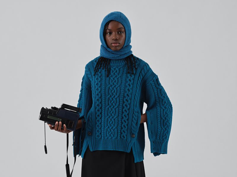 Portrait of a young Black woman wearing a teal sweater and hood, holding a black video camera against a plain white backdrop.