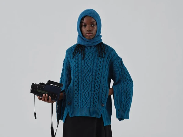 Portrait of a young Black woman wearing a teal sweater and hood, holding a black video camera against a plain white backdrop.