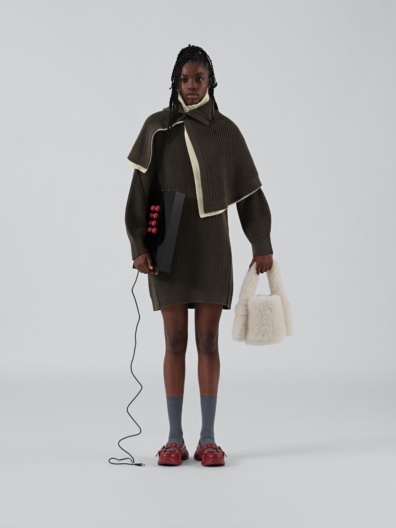Full-length shot of a black female model in a knit dress with a capelet, red shoes, and a black box with red knobs, in a bright studio.