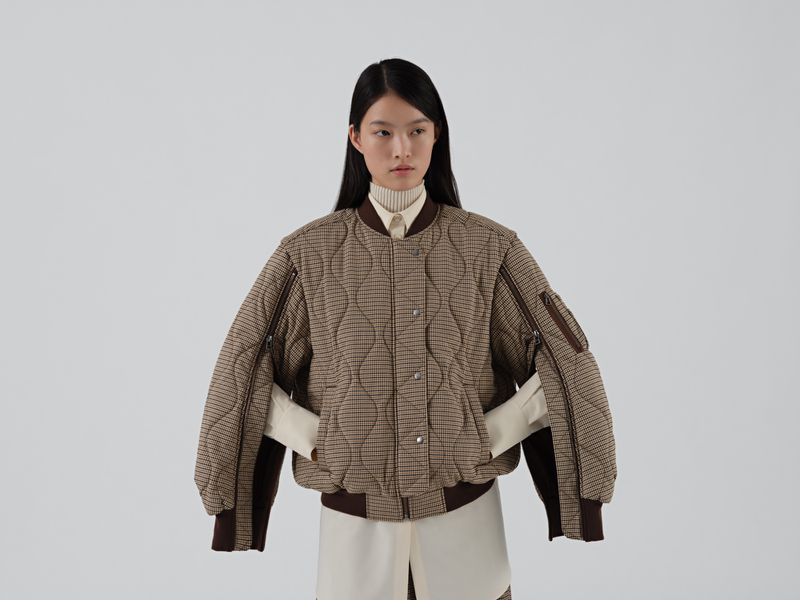 Model wearing a brown quilted bomber jacket with arm zippers over a cream shirt against a white seamless background