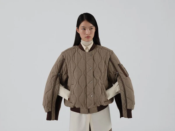 Model wearing a brown quilted bomber jacket with arm zippers over a cream shirt against a white seamless background