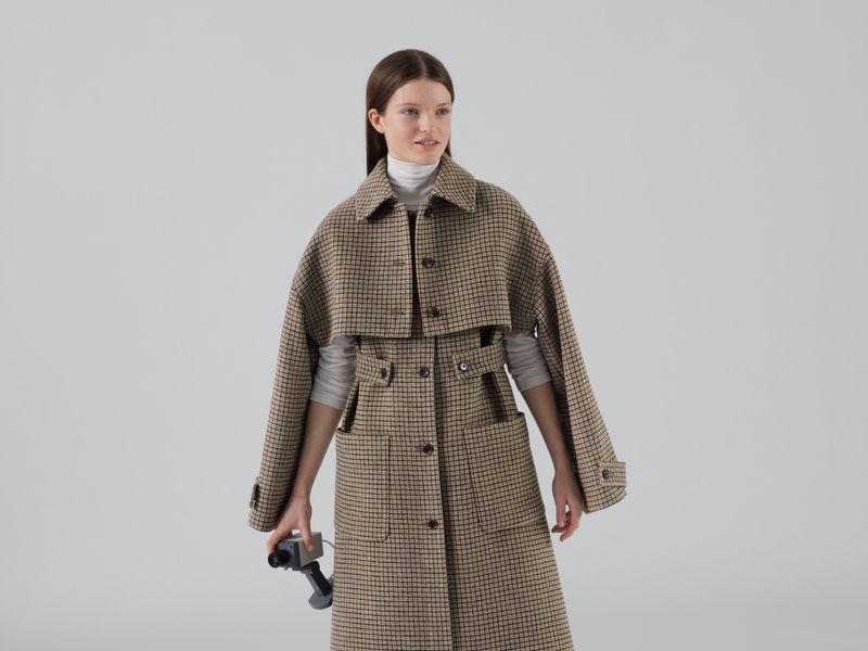 A female model wears a brown plaid overcoat, holding an old handheld video camera against a light gray seamless background.