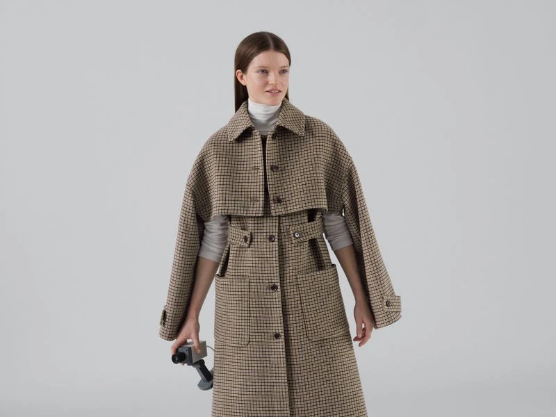 A female model wears a brown plaid overcoat, holding an old handheld video camera against a light gray seamless background.