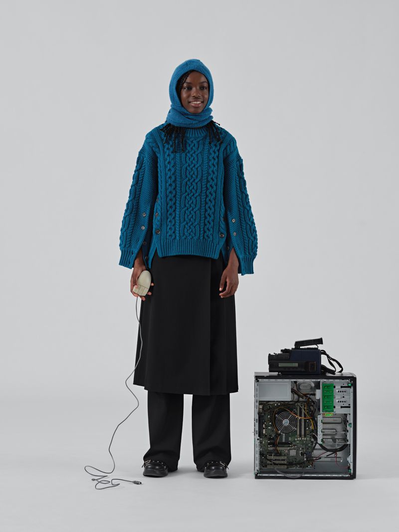 A black woman in a teal knit balaclava and sweater stands in a light grey studio holding a computer mouse next to a desktop computer.