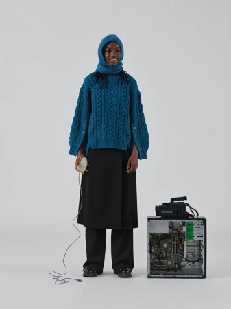A black woman in a teal knit balaclava and sweater stands in a light grey studio holding a computer mouse next to a desktop computer.