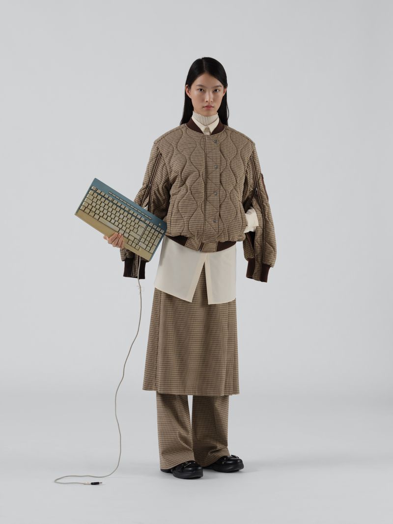 Full-shot of a young adult woman in a brown quilted bomber jacket, skirt, and pants, holding a keyboard in a white photo studio.